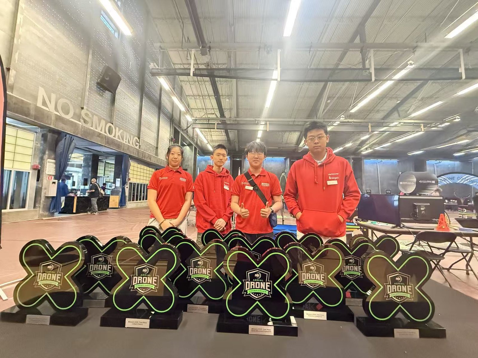 Four students in red team tops standing behind rows of Aerial Drone Championship trophies on a table