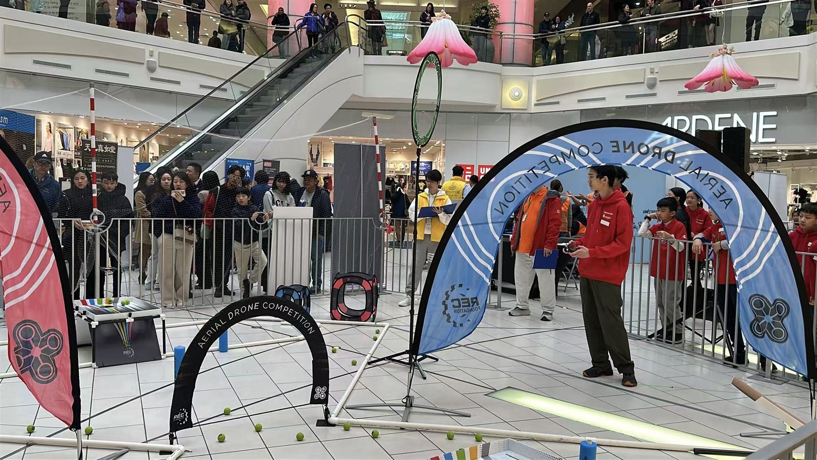 A shopping-centre atrium set up as a drone competition course: hoop and arch gates on the tiled floor, team tables behind barriers, and spectators watching from the balconies