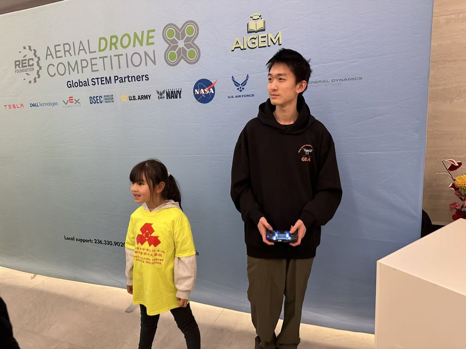 Terry holding a transmitter beside a young student in a yellow Chinese Cultural Centre shirt, in front of an Aerial Drone Competition backdrop