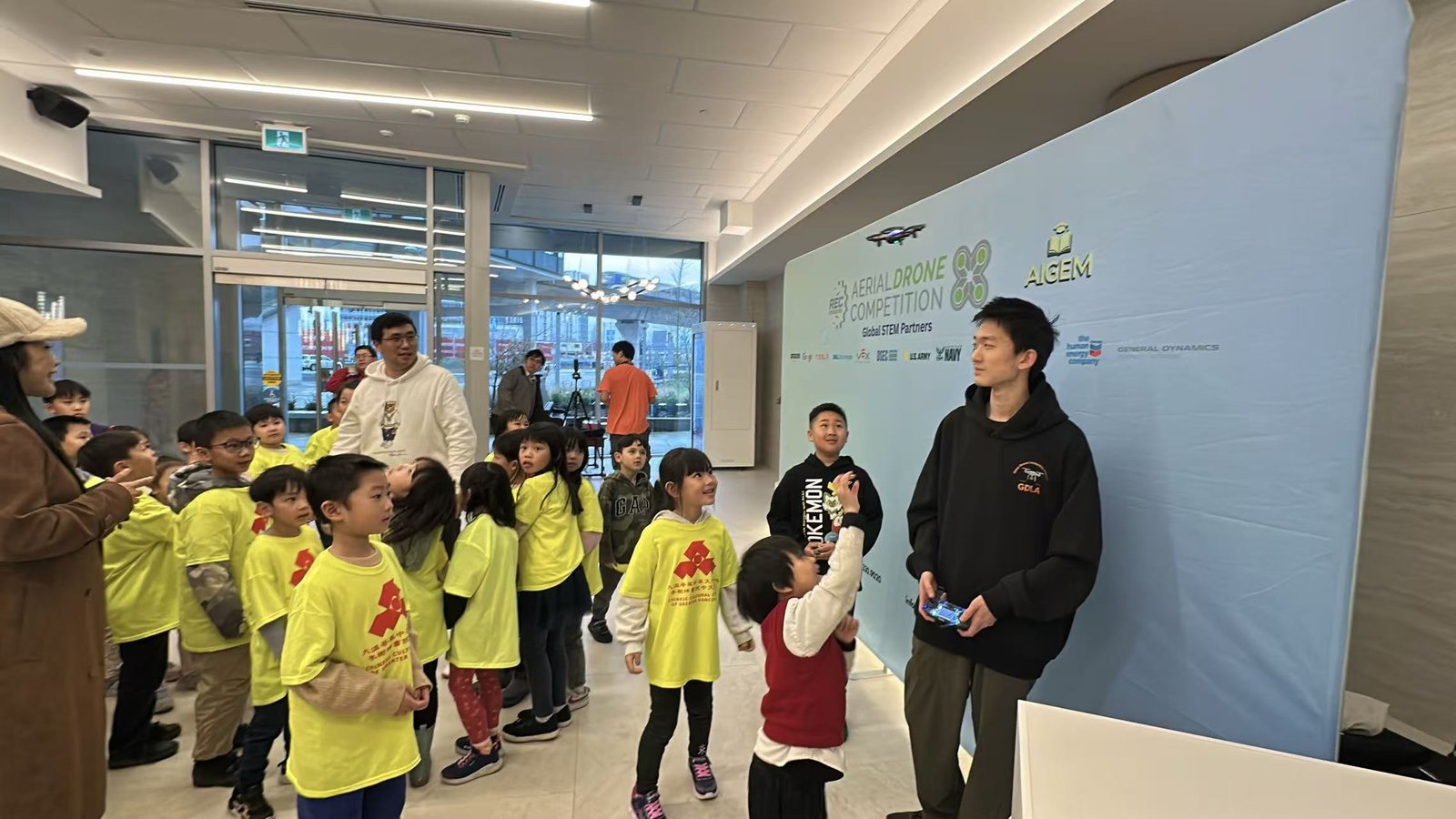 Children in yellow Chinese Cultural Centre shirts gathered around Terry, who is holding a transmitter, in front of an Aerial Drone Competition backdrop